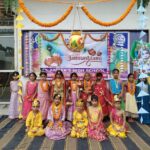 Group of indian children in colorful traditional outfits posing for a janmashtami celebration in front of a decorated school backdrop with marigold garlands and paper banners